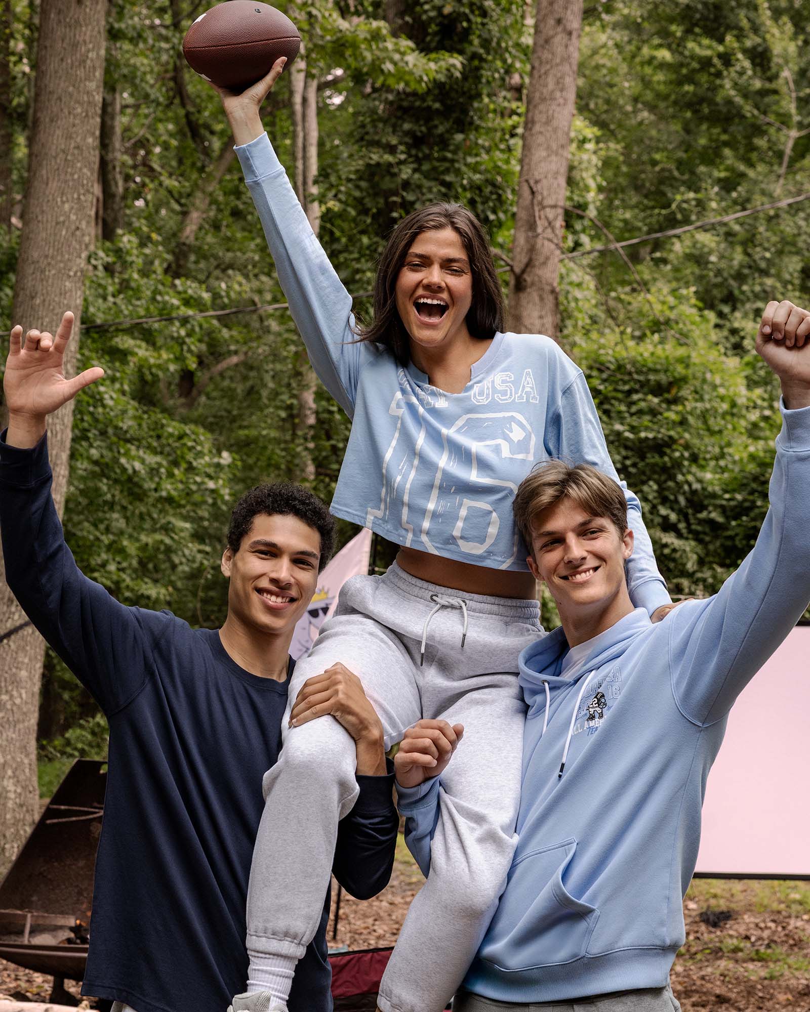 Three people celebrating outdoors with a football, one person is lifted in the air.