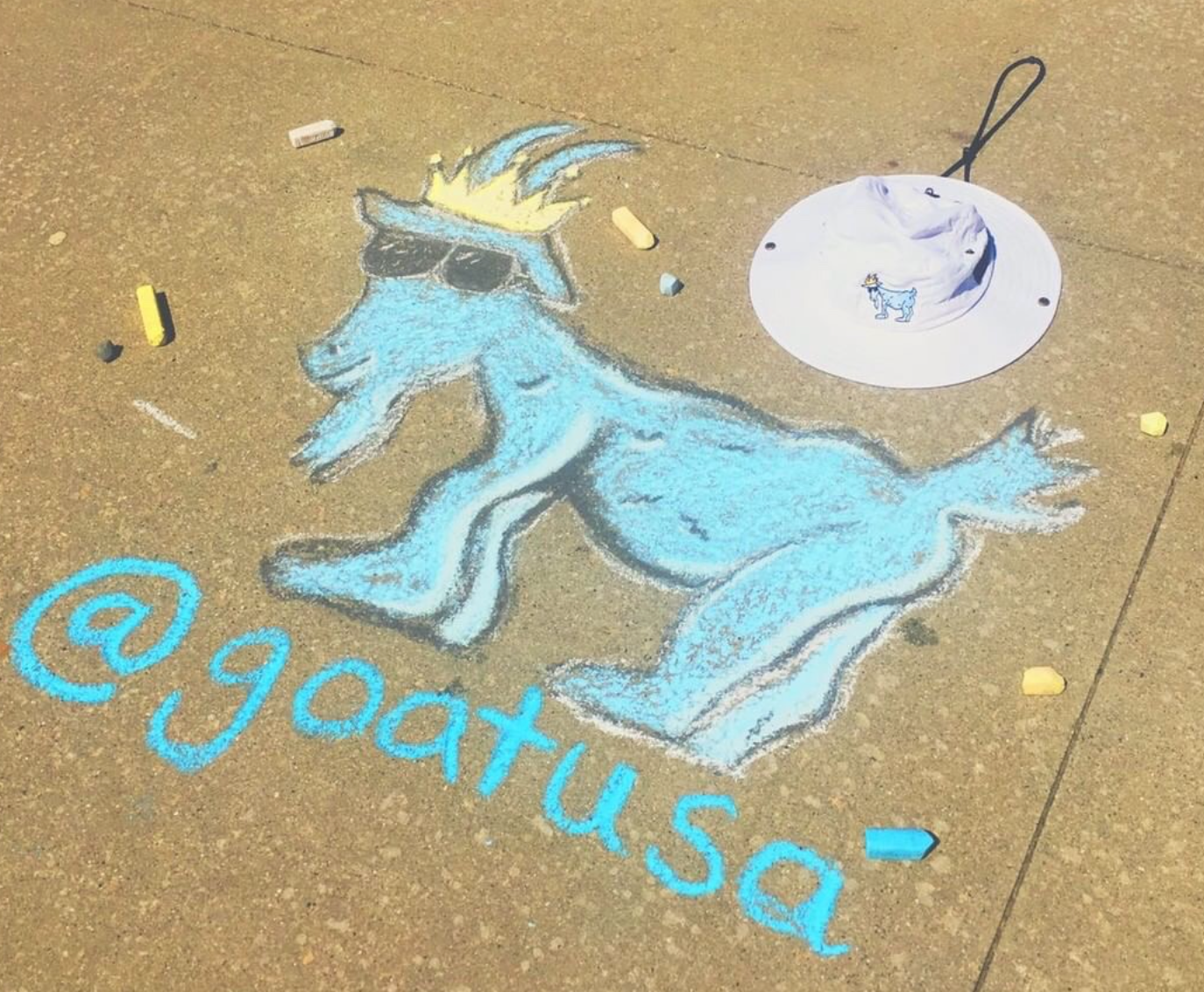 Chalk drawing on sidewalk of GOAT USA goat logo with white bucket hat in the frame