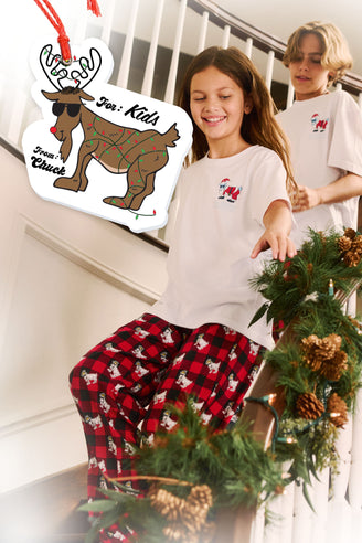 Child wearing red and black checkered pajama pants with a reindeer design, standing on a staircase with a Christmas wreath.