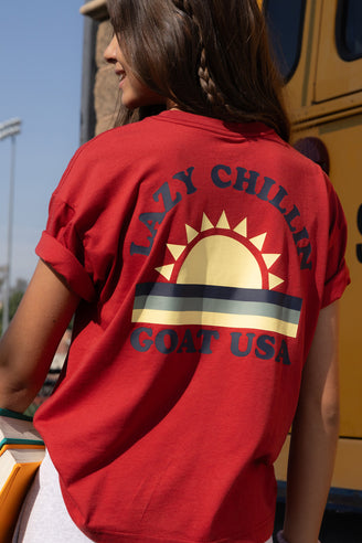 A woman viewed from behind wearing a red graphic t-shirt with a large 'Lazy Chillin' Goat USA' sun design on the back, standing outdoors next to a yellow bus.