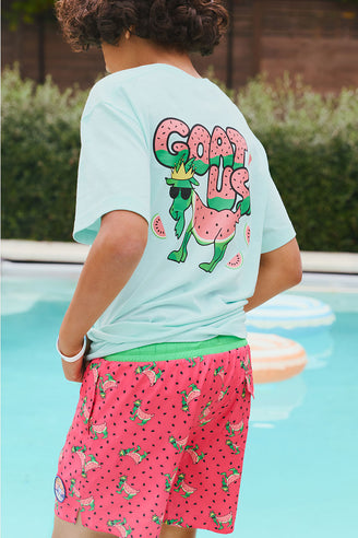 Child wearing a light green t-shirt with a graphic design and pink shorts with watermelon pattern by a pool.