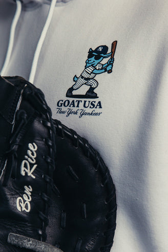 Close-up of a light gray hoodie with a black baseball glove in the foreground and a printed graphic on the chest showing a cartoon goat in a New York Yankees baseball uniform with the text 'GOAT USA New York Yankees'.