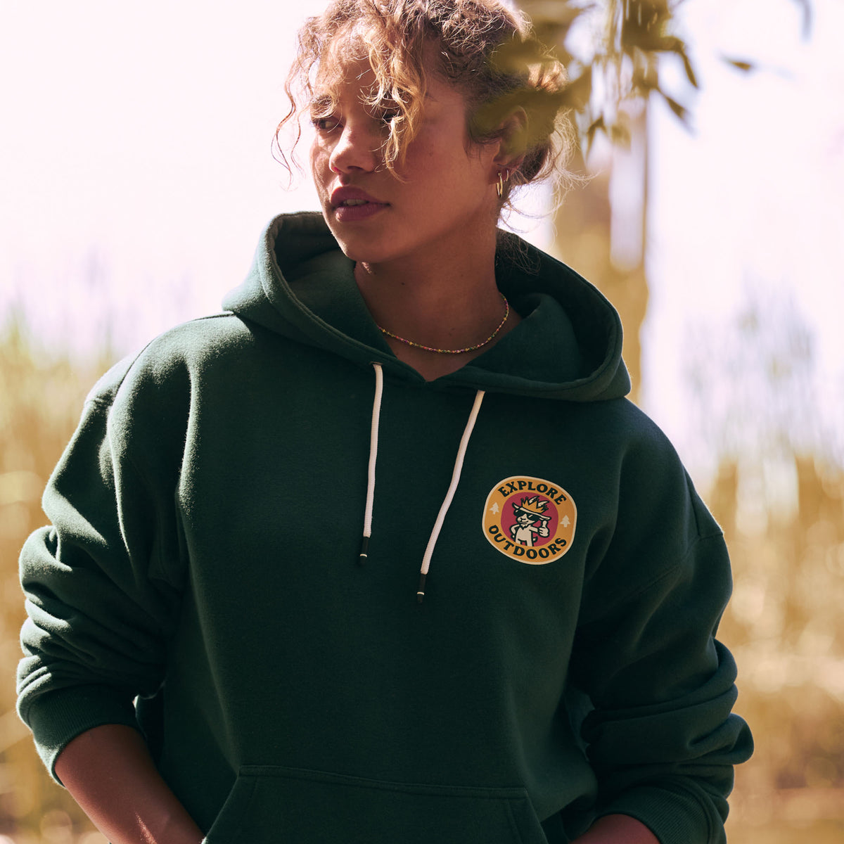 A woman wearing a dark green pullover hoodie with white drawstrings and a circular 'Explore Outdoors' logo patch on the left chest, photographed outdoors in warm natural light.