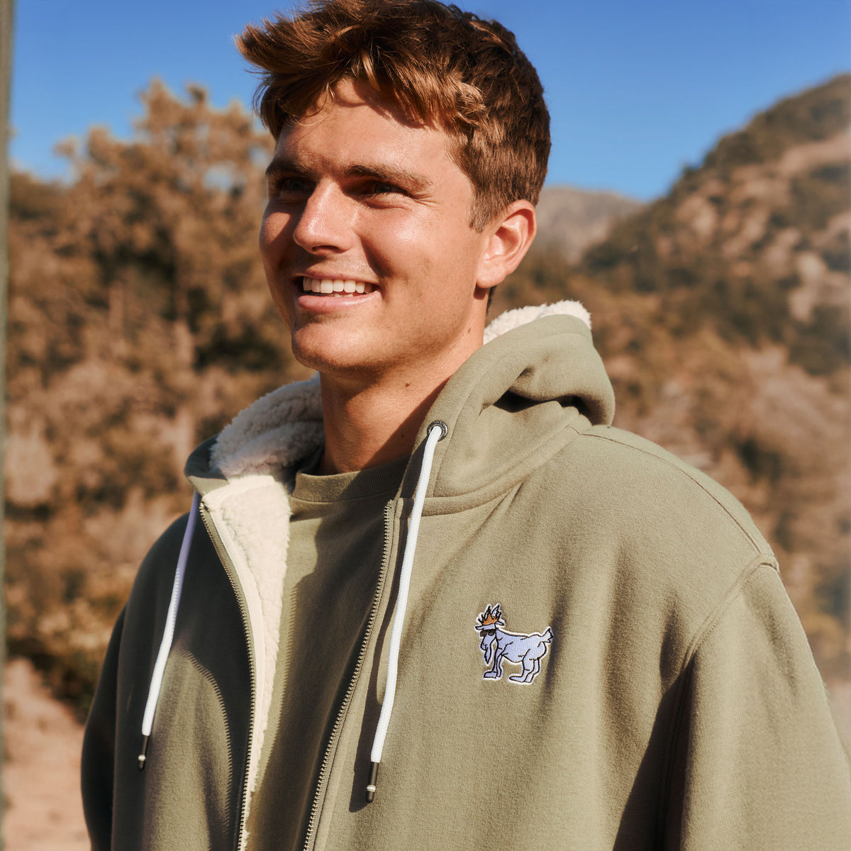 A smiling young man outdoors wearing an olive green zip-up hoodie with a white sherpa lining and a small embroidered goat logo on the chest, set against a blurred mountain background.