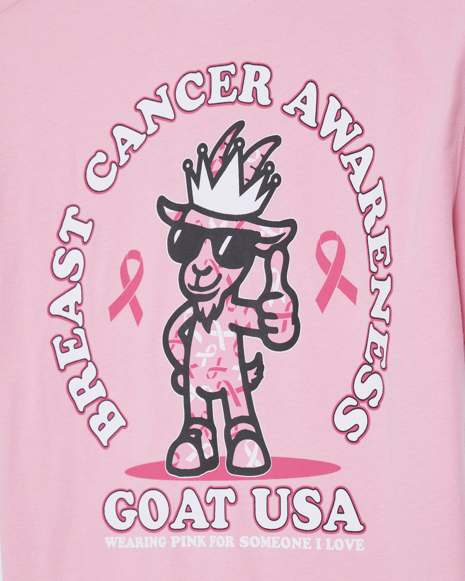 A close-up of a pink graphic t-shirt featuring a cartoon goat character wearing sunglasses and a crown, surrounded by the text 'BREAST CANCER AWARENESS' and 'GOAT USA'.