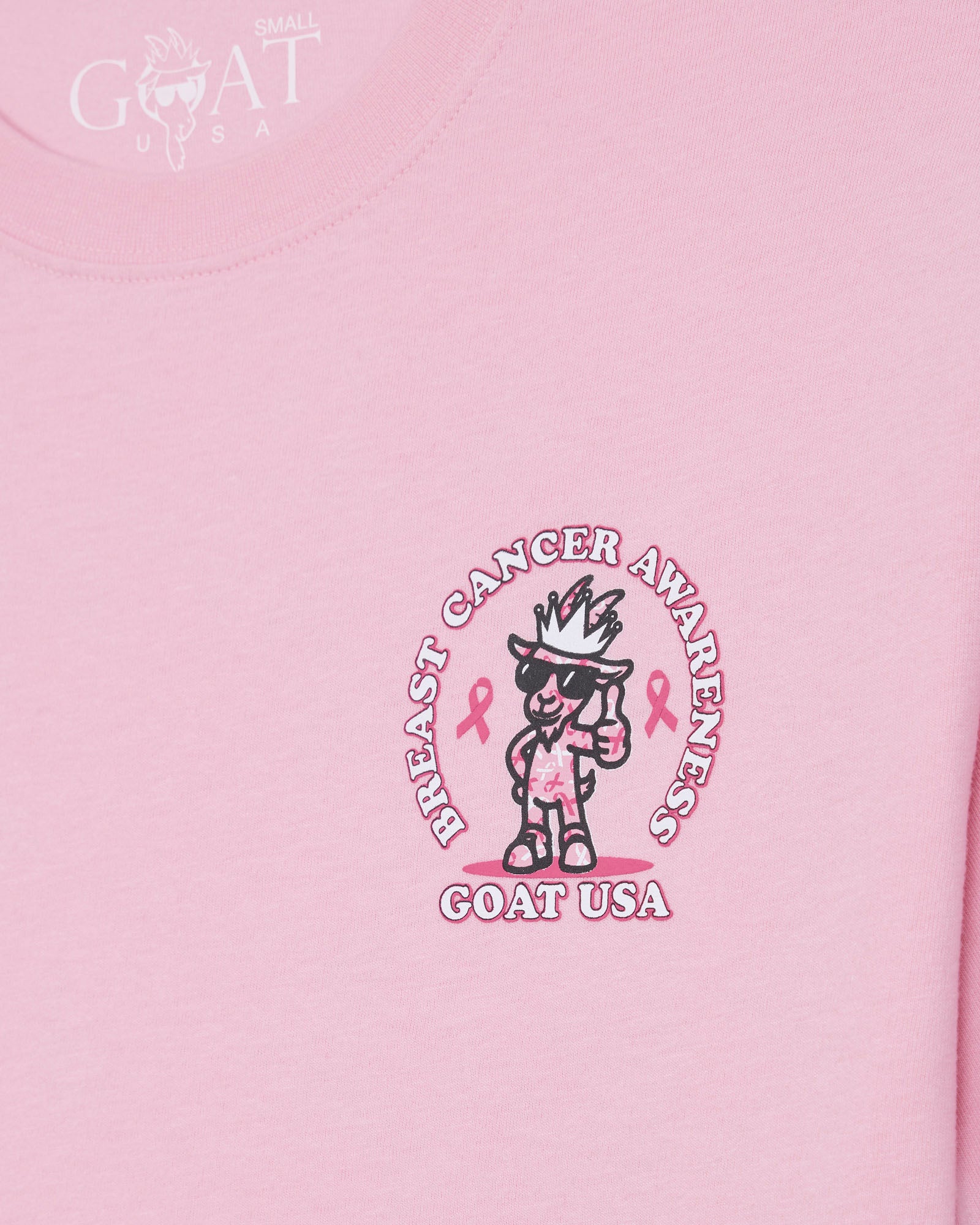 A close-up of a light pink t-shirt featuring a central graphic for Breast Cancer Awareness. The graphic shows a cartoon goat character wearing a crown and sunglasses, with pink awareness ribbons and the text 'BREAST CANCER AWARENESS' and 'GOAT USA' written around it.