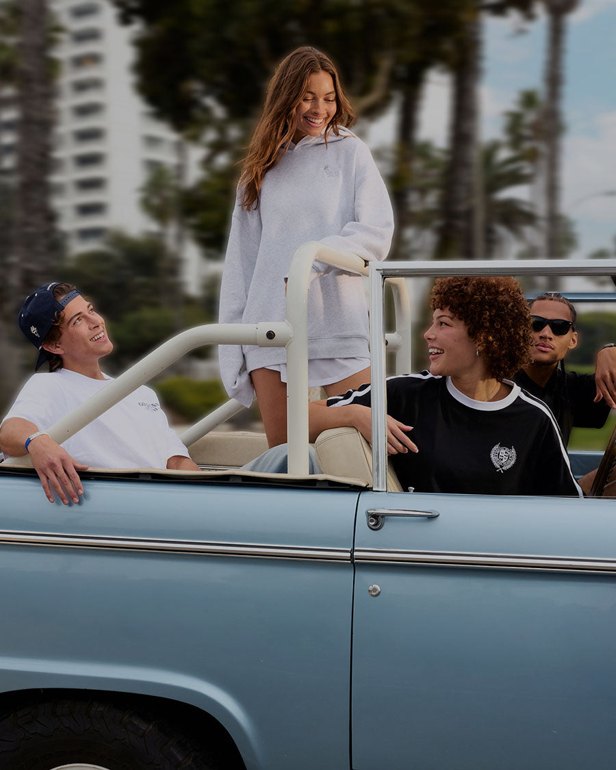 Group of people in a vintage convertible car with palm trees and buildings in the background