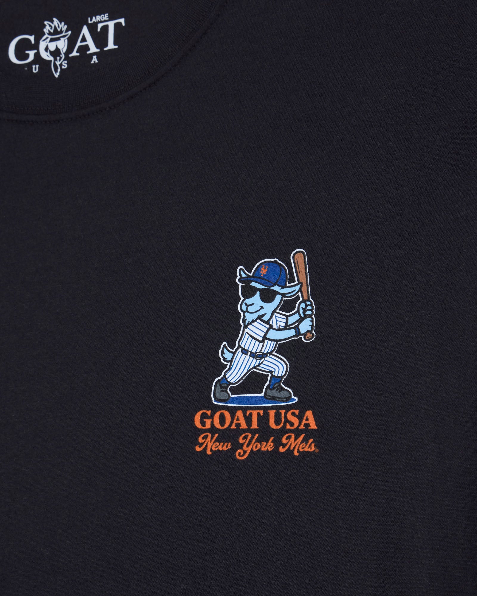 Close-up of a black t-shirt featuring a central graphic of a blue and white goat mascot holding a baseball bat, with 'GOAT USA New York Mets' below in orange. The top left corner shows a white 'GOAT USA LARGE' brand print.