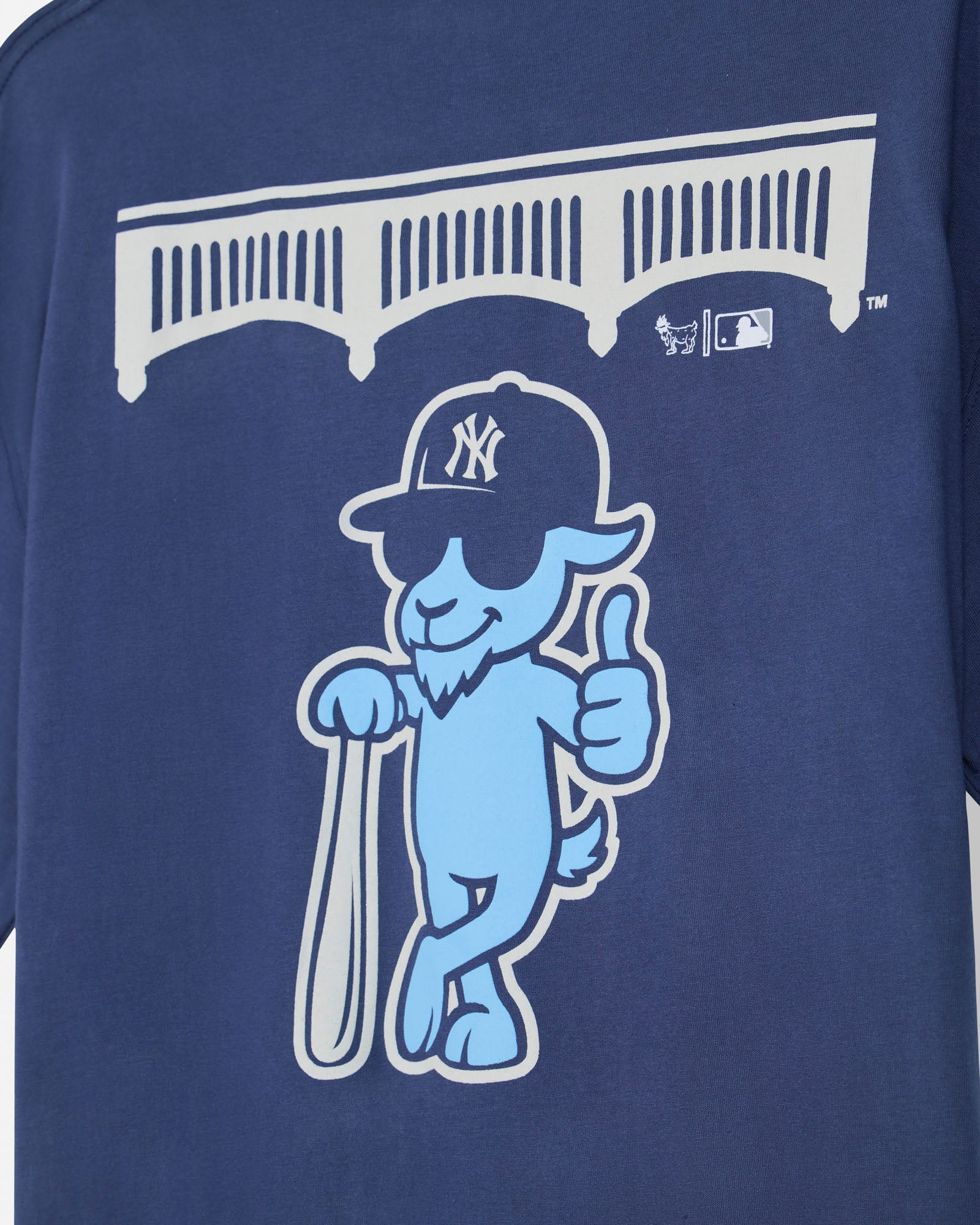 Close-up of a navy blue T-shirt featuring a light blue cartoon goat mascot wearing a New York Yankees cap, holding a bat, and giving a thumbs-up, with a white stadium graphic above.