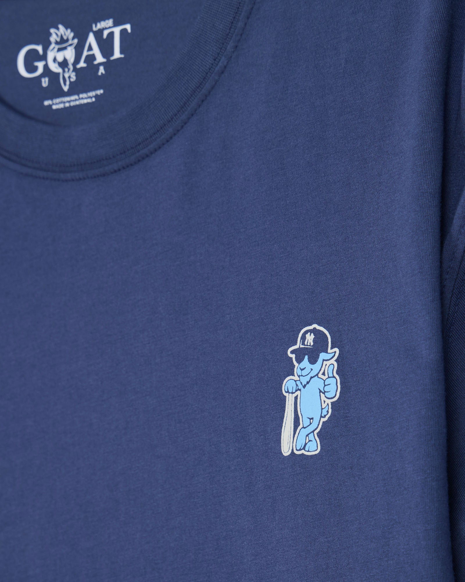 Close-up of a navy blue t-shirt featuring a small embroidered light blue and white cartoon character on the left chest, with the Goat USA brand logo printed inside the collar.