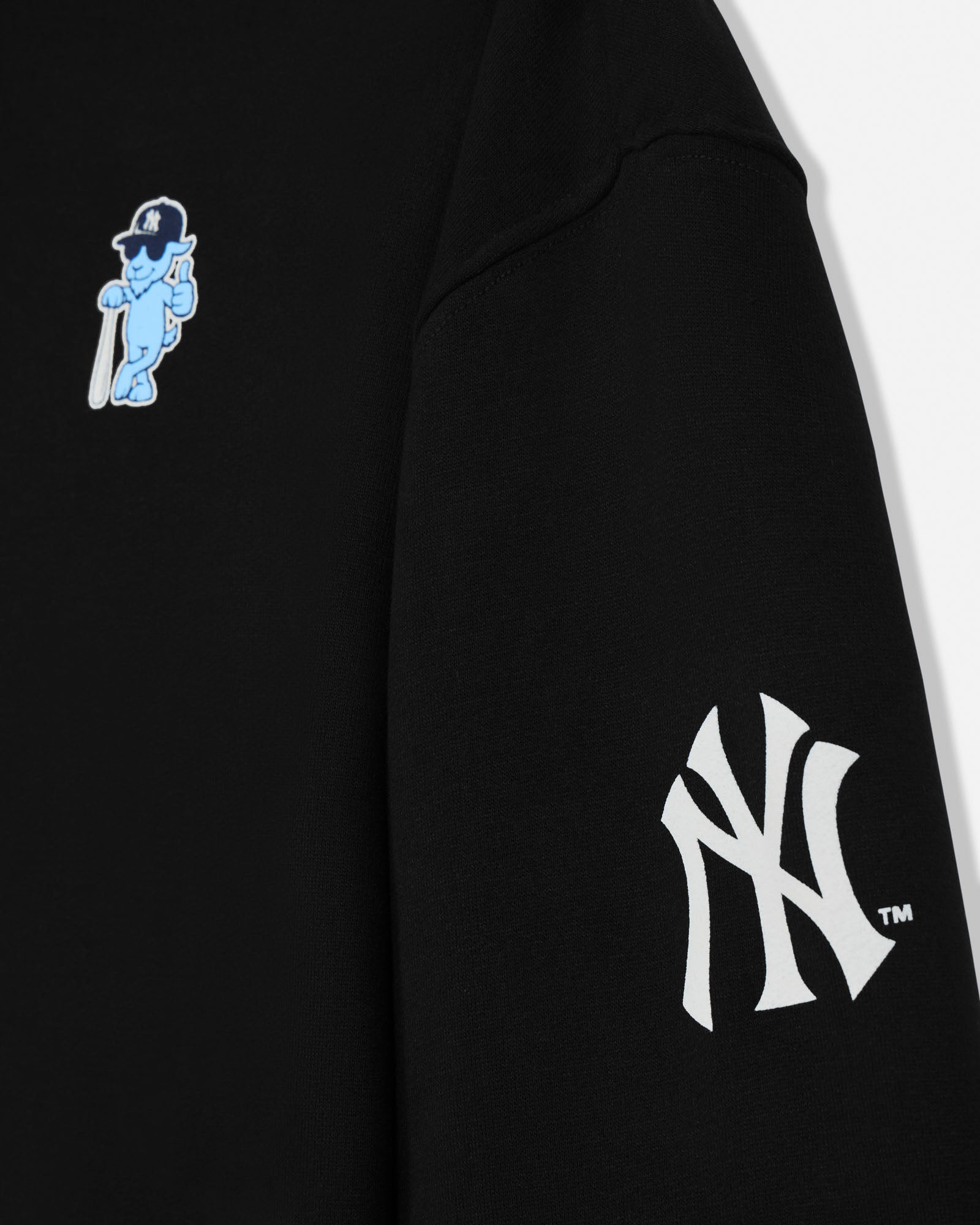 Close-up of a black sweatshirt sleeve featuring a white New York Yankees interlocking NY logo on the lower arm and a small light blue cartoon character patch on the upper sleeve.