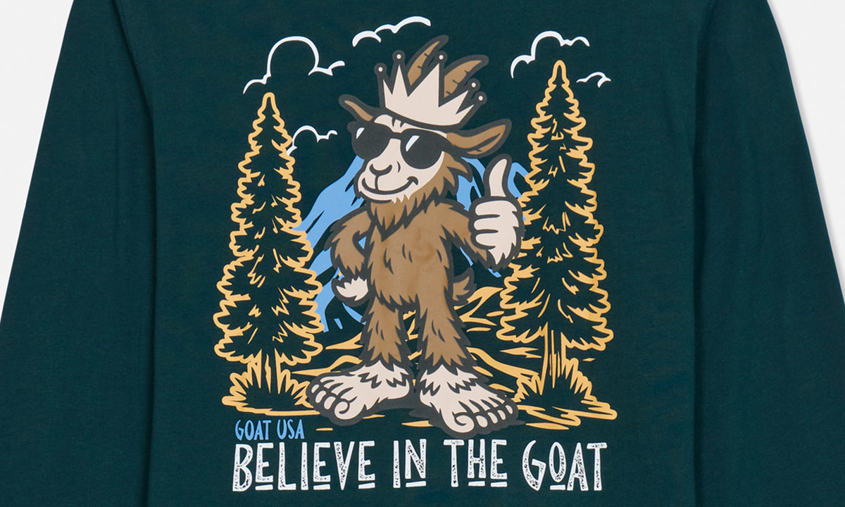 A close-up of a dark teal long-sleeve shirt featuring a graphic of a brown cartoon goat wearing sunglasses and a crown, standing between two pine trees with the text 'GOAT USA BELIEVE IN THE GOAT' below.