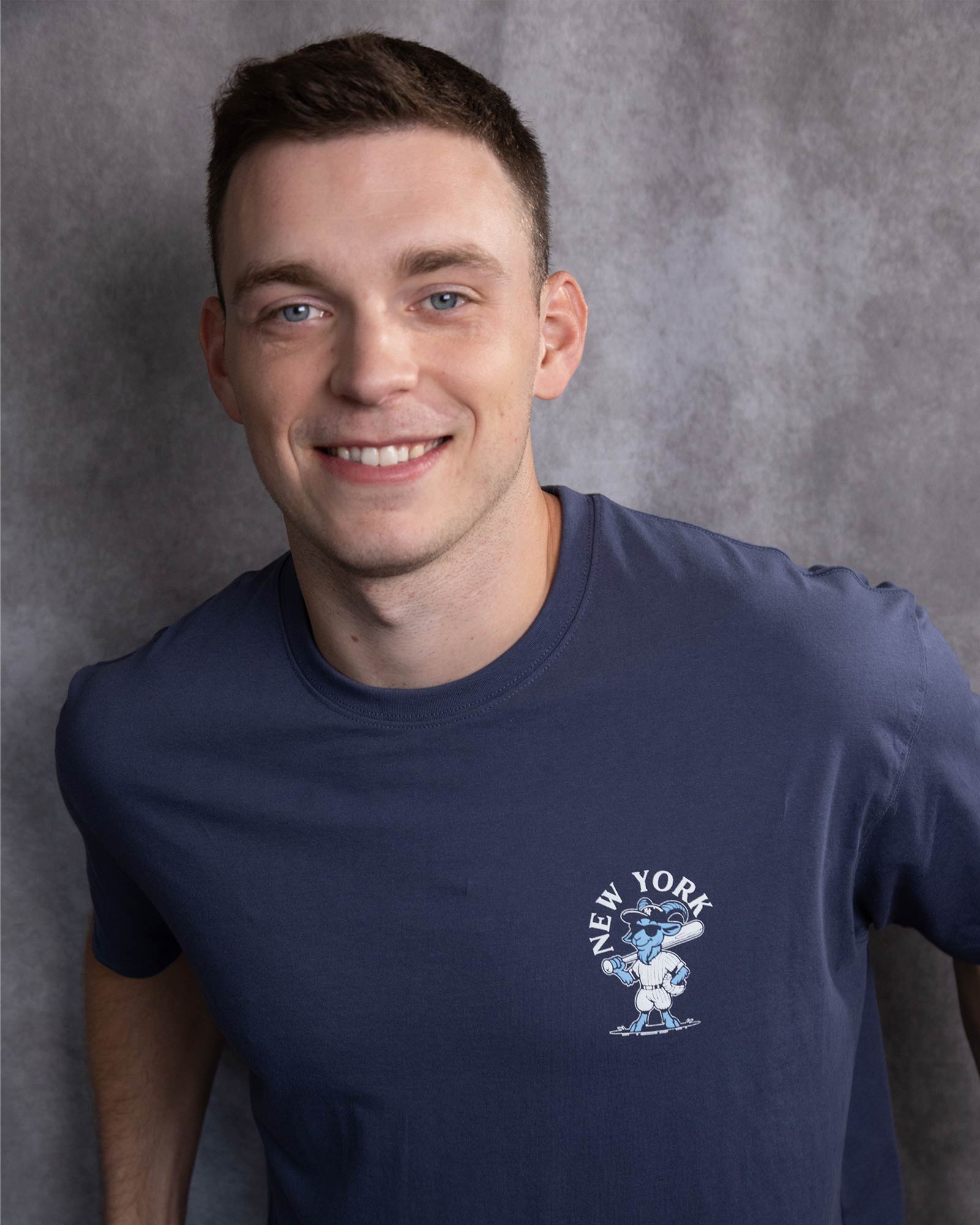 A smiling man wearing a navy blue crewneck t-shirt with a white and blue New York Yankees graphic on the left chest, set against a textured gray studio background.