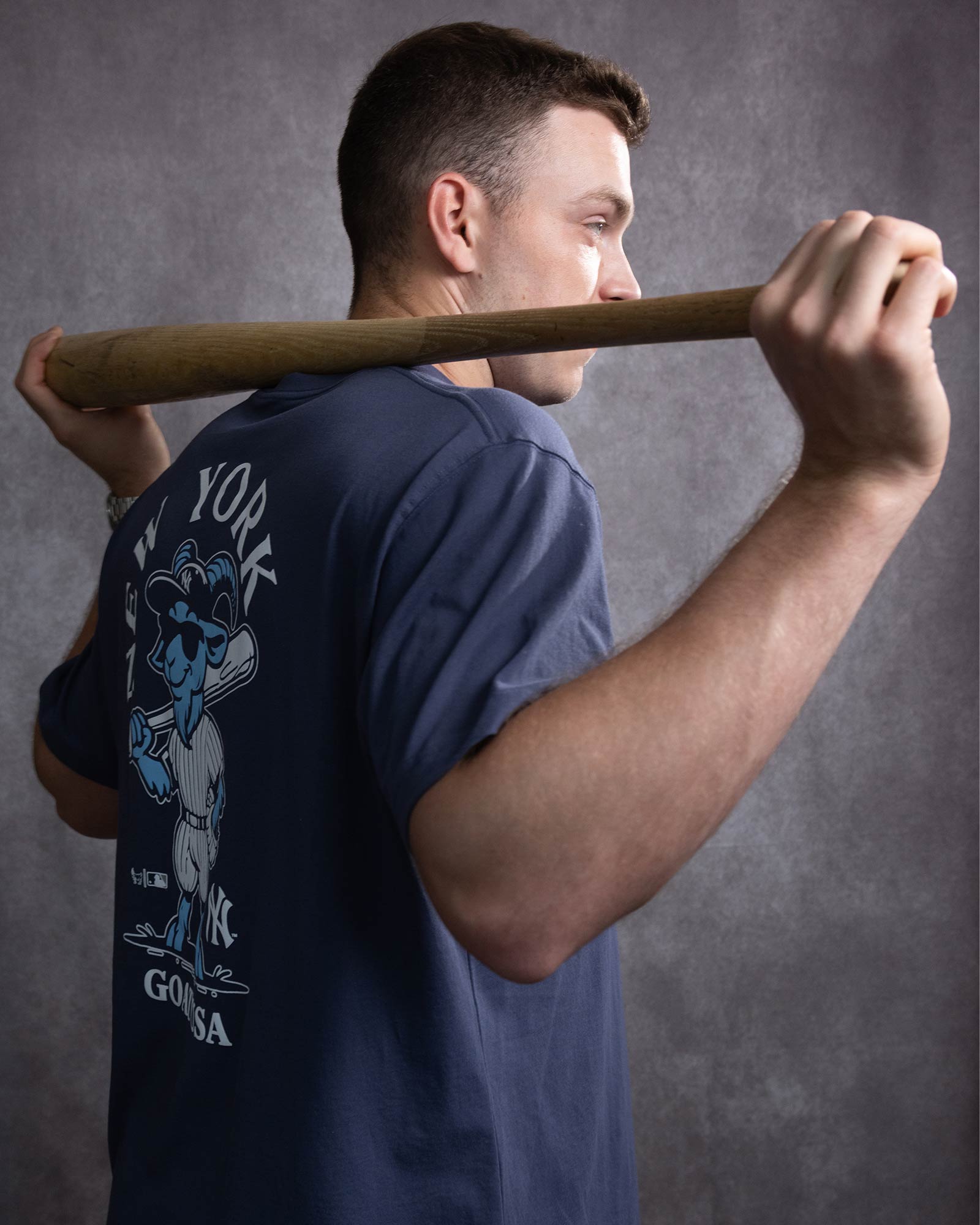 A man holding a baseball bat across his shoulders against a textured gray studio background