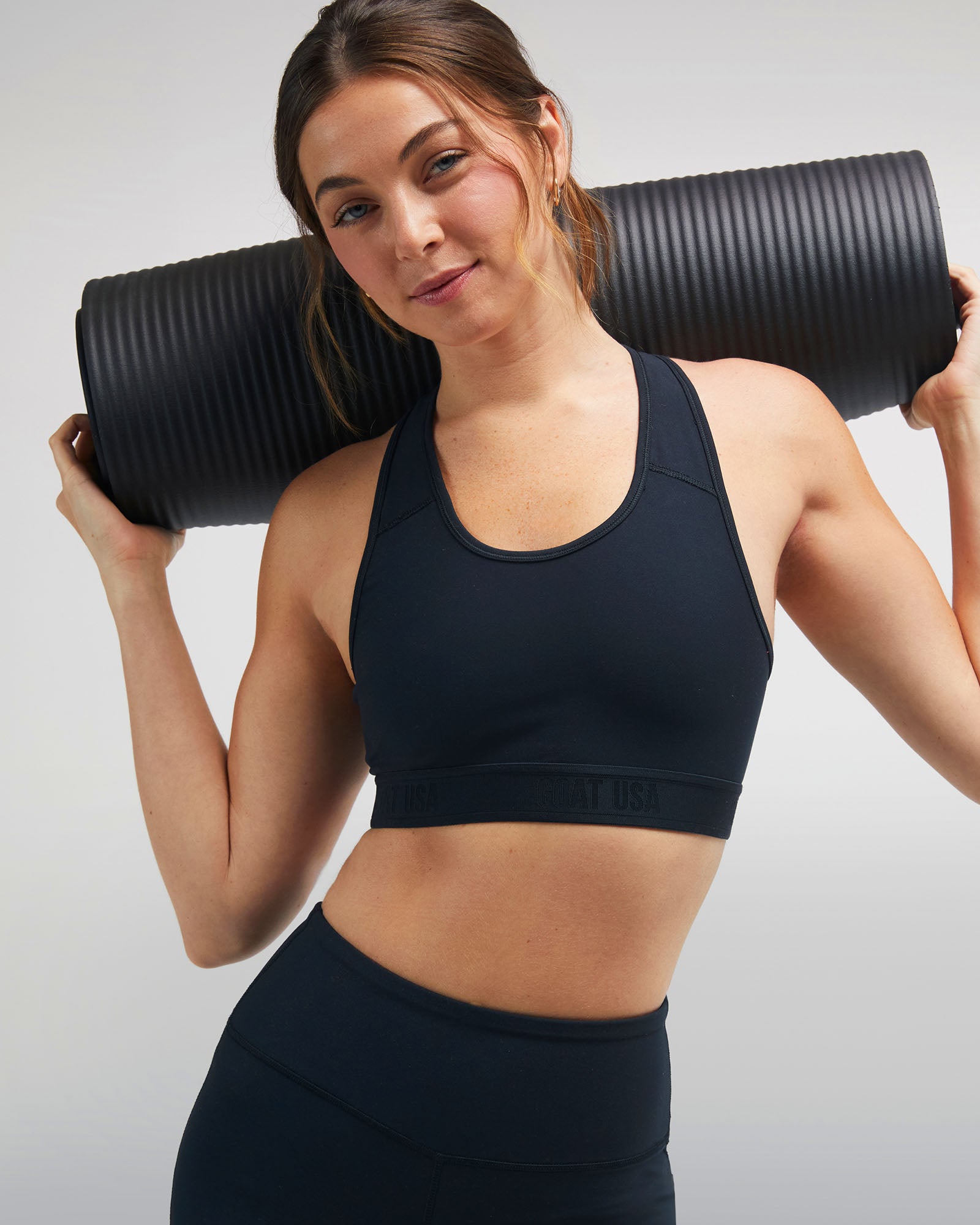 Woman holding a black rolled up yoga mat against a plain background