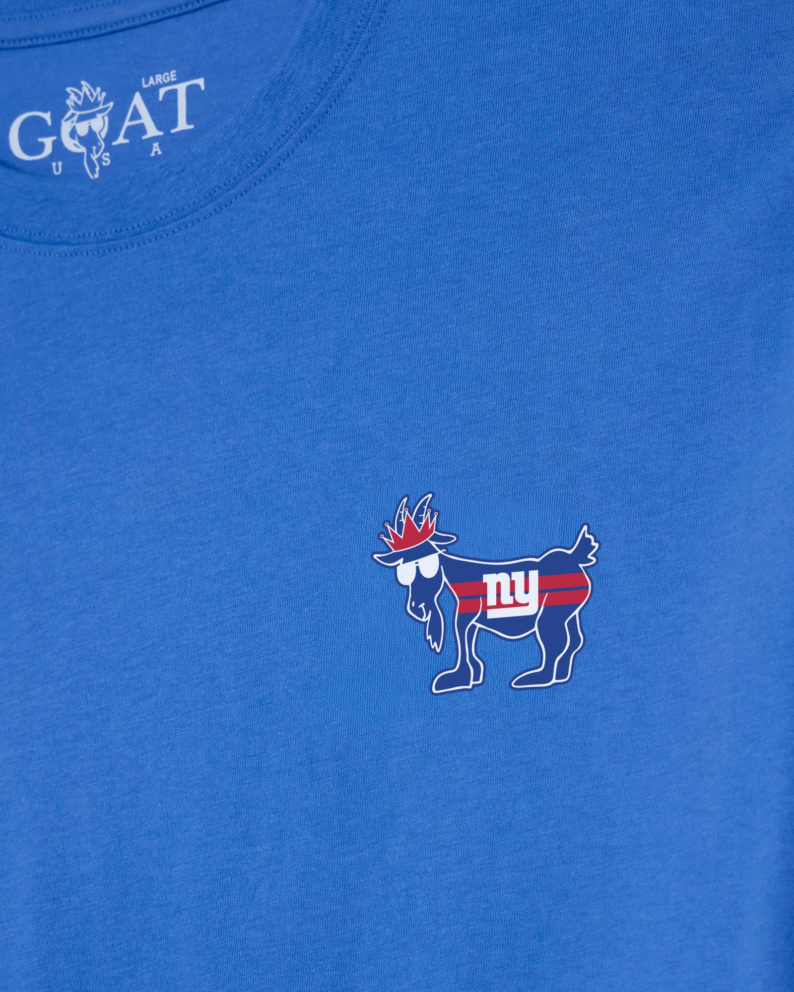 A close-up of a royal blue t-shirt featuring a small New York Giants goat mascot graphic on the left chest with red, white, and blue details, and the white 'GOAT USA' brand logo printed inside the collar.