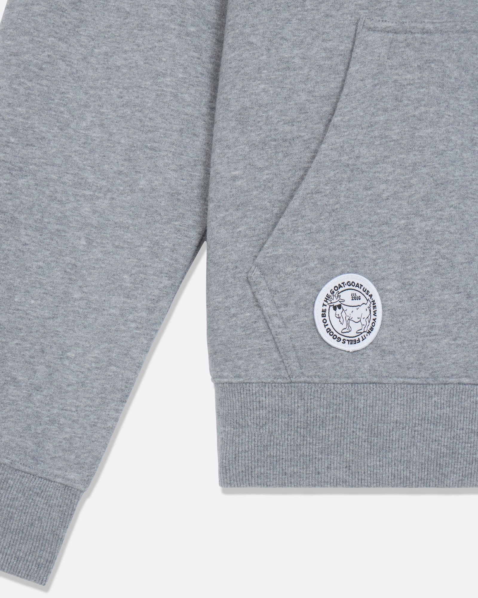 Close-up of a heather gray sweatshirt sleeve with a ribbed cuff and a circular white embroidered patch featuring a goat.