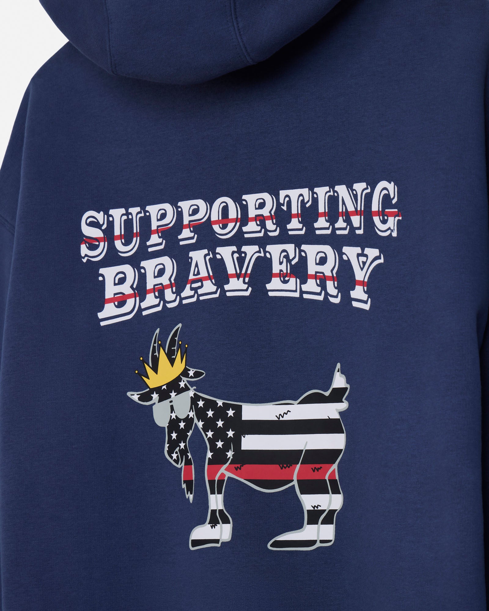 Close-up of the back of a navy blue hoodie featuring a graphic design with the words 'SUPPORTING BRAVERY' in white outlined letters with red strike-through lines, and a goat illustration filled with an American flag pattern and a gold crown.