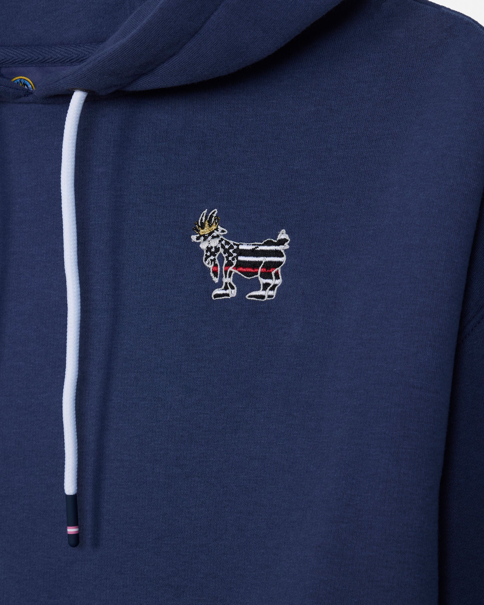 Close-up of a navy blue hoodie featuring a white drawstring and a small embroidered goat graphic on the chest with red, white, and black stripes and a gold crown.