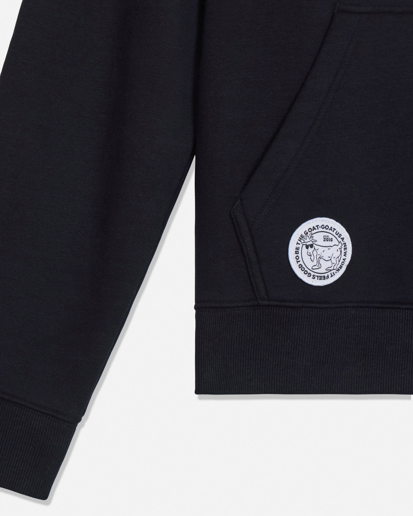 Close-up of a black crewneck sweatshirt showing the left sleeve and chest area, featuring a circular white embroidered logo patch on the pocket.