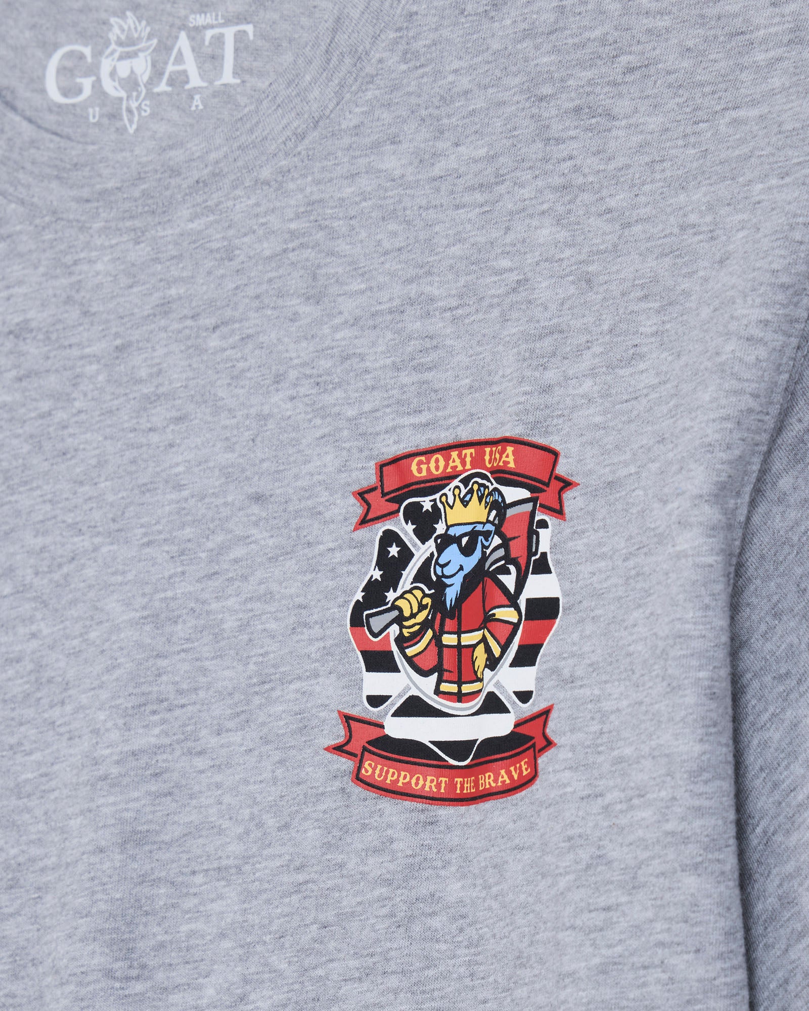 Close-up of a heather gray t-shirt featuring a colorful graphic on the chest. The graphic displays a cartoon blue character wearing a red and gold crown, flanked by red banners reading 'GOAT USA' and 'SUPPORT THE BRAVE'.