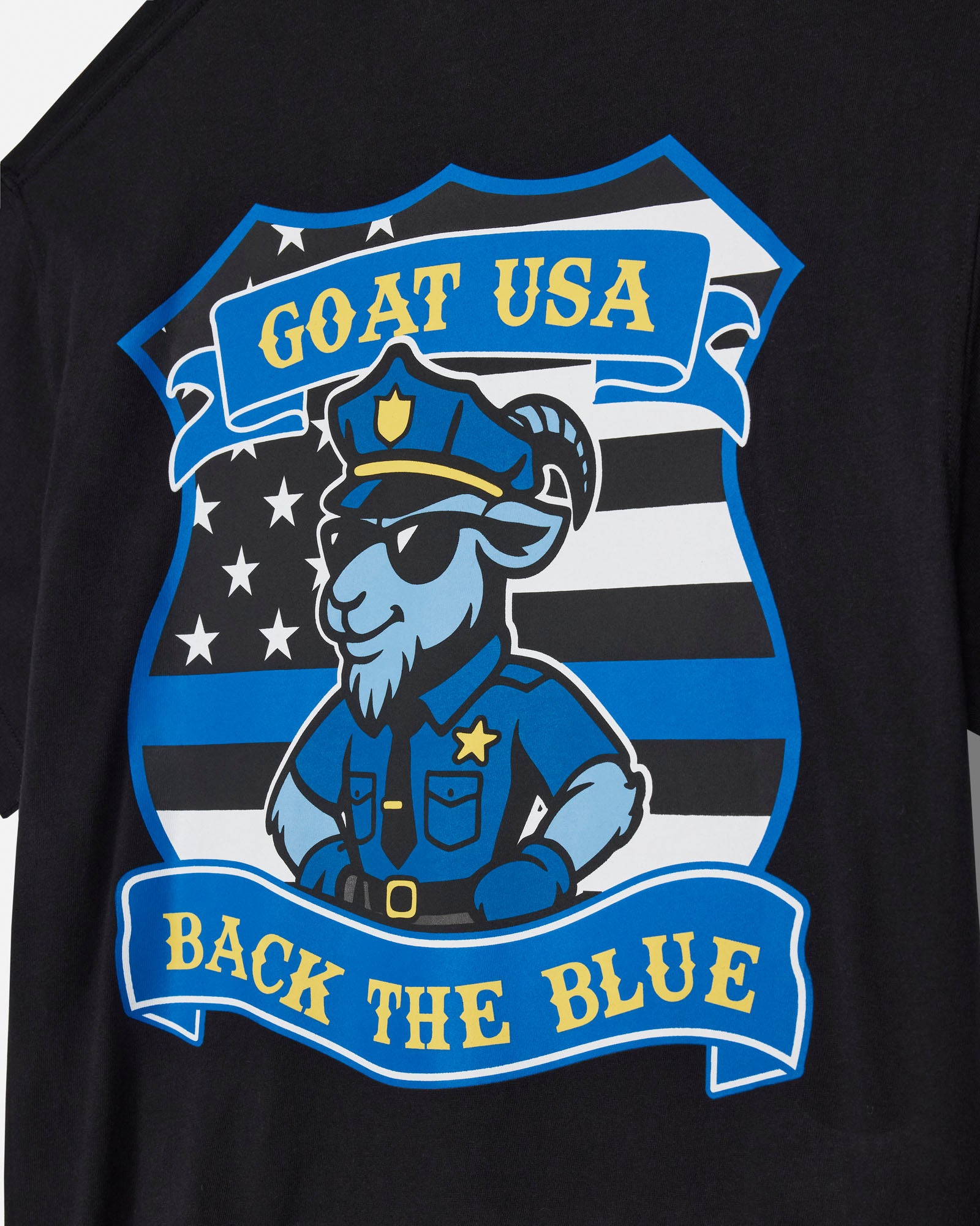 Close-up of a black T-shirt featuring a large graphic print of a cartoon goat in a police uniform with the text 'GOAT USA' and 'BACK THE BLUE' on blue banners, set against a black and white striped shield design.