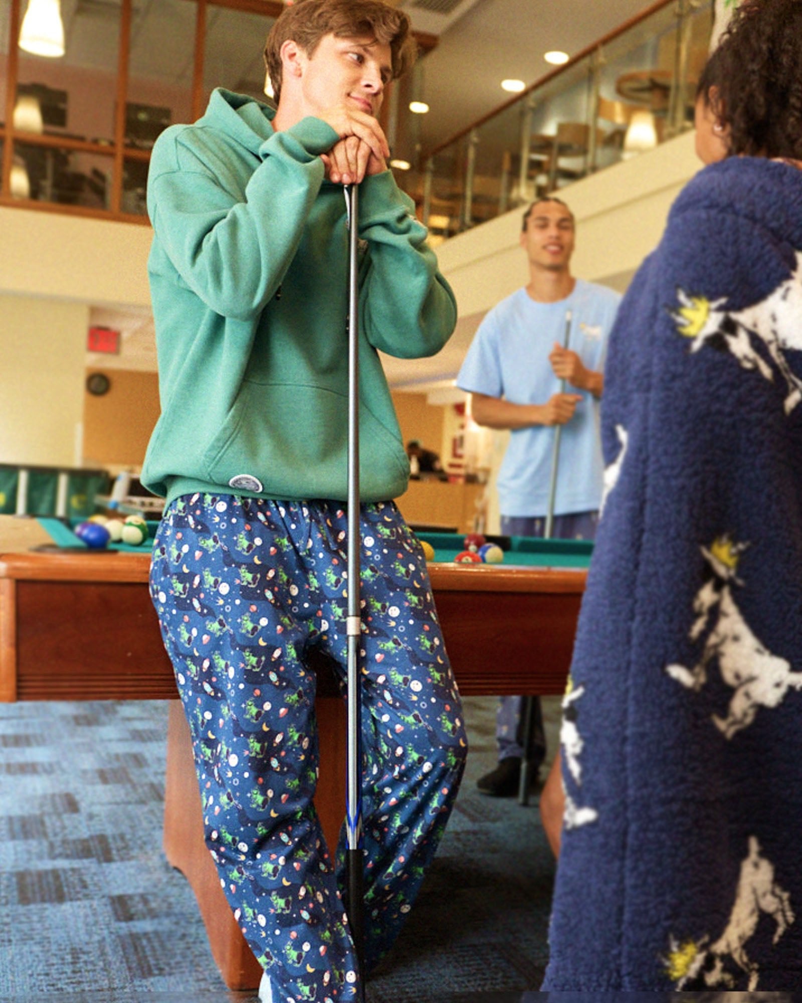 Person in green hoodie and patterned pants playing pool with another person.