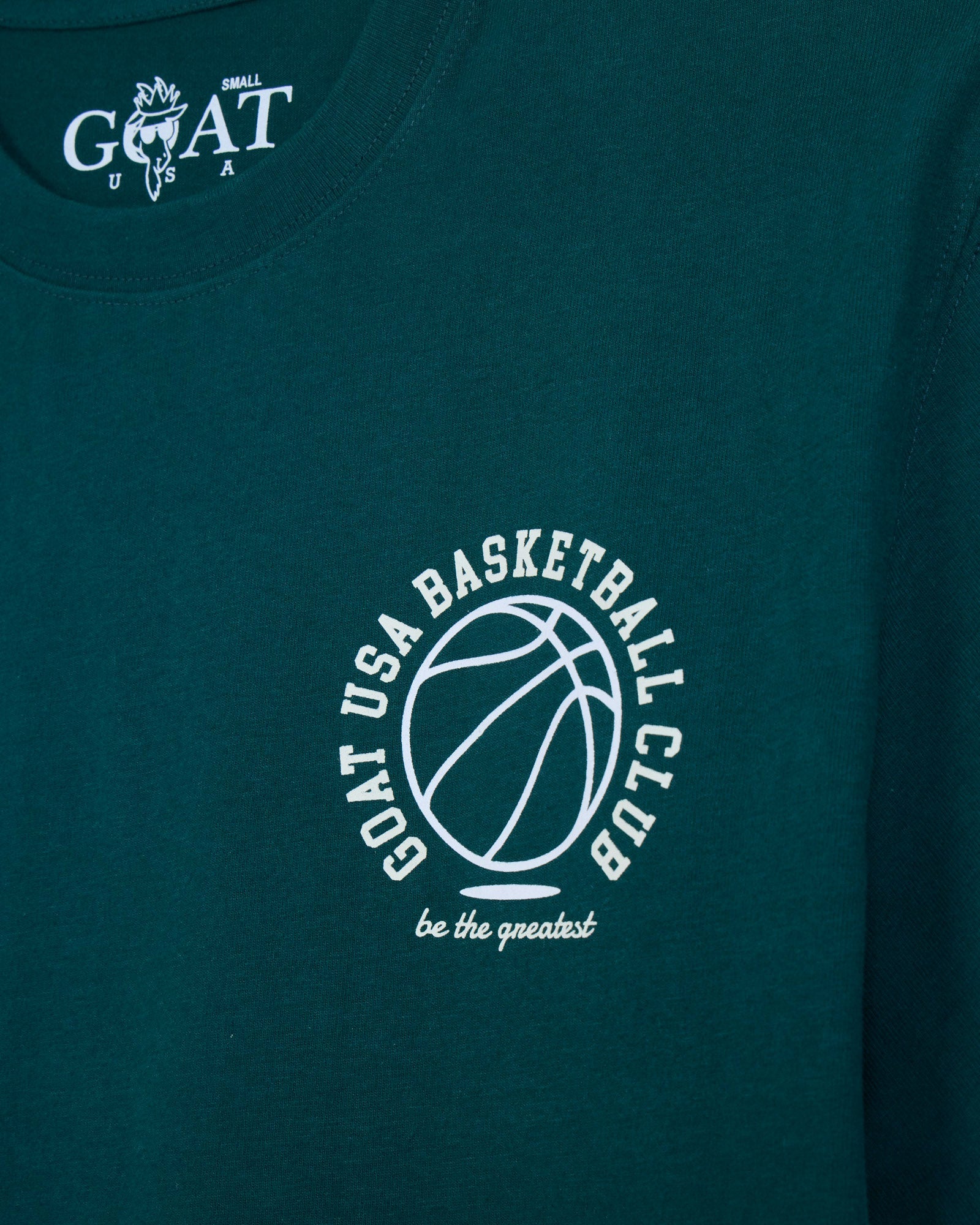 Close-up of a dark teal t-shirt featuring a white circular graphic on the chest with a basketball outline and the text 'GOAT USA BASKETBALL CLUB be the greatest'. The inside collar shows a white 'GOAT' logo.