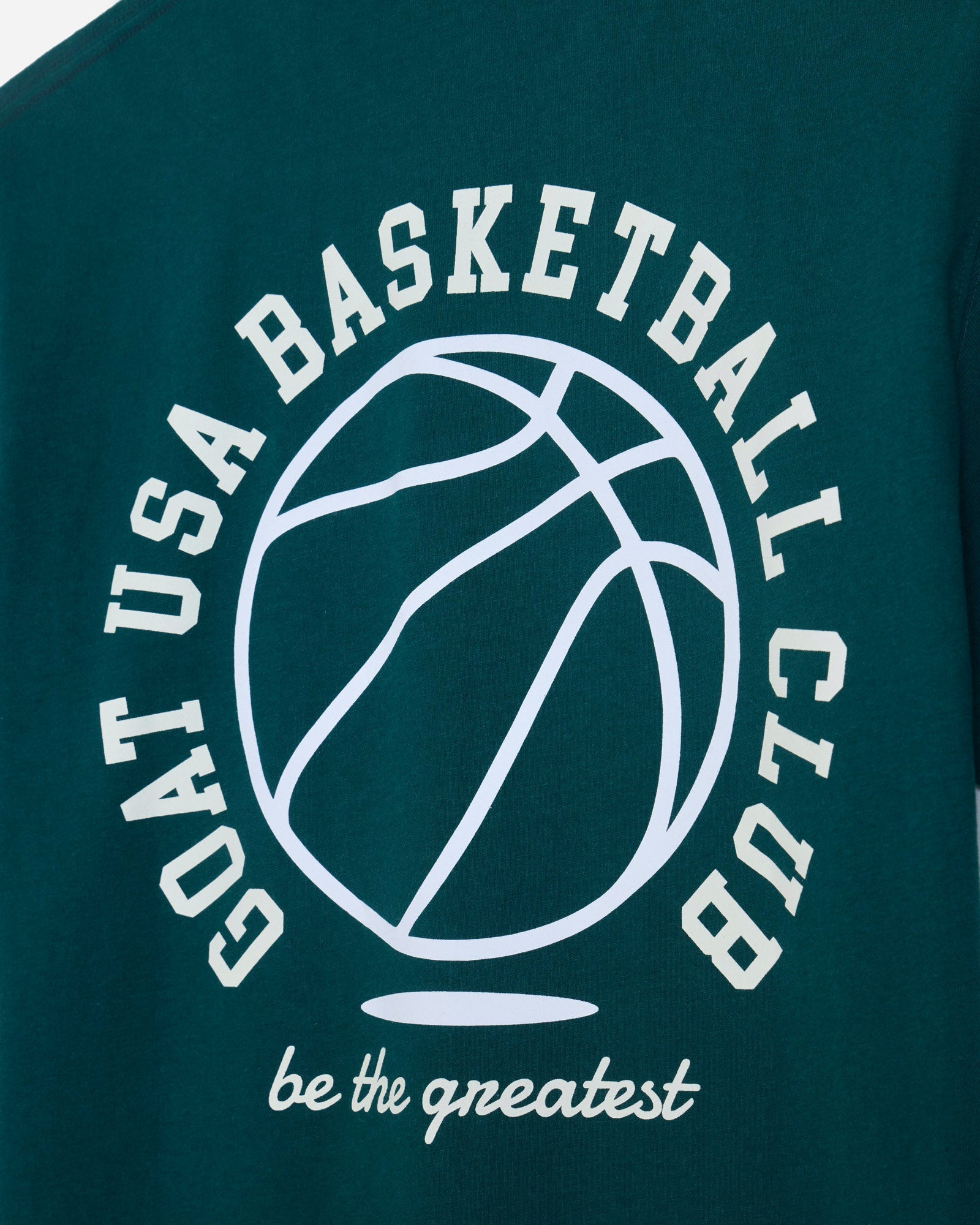 Close-up of a dark teal t-shirt featuring a white graphic print of a basketball and the text 'GOAT USA BASKETBALL CLUB' and 'be the greatest'.