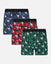 Three pairs of men's boxer briefs in holiday-themed plaid patterns, displayed in a staggered stack on a light gray background. The top pair is navy blue and black with white goat prints, the middle pair is red and black with white goat prints, and the bottom pair is green and black with white goat prints.