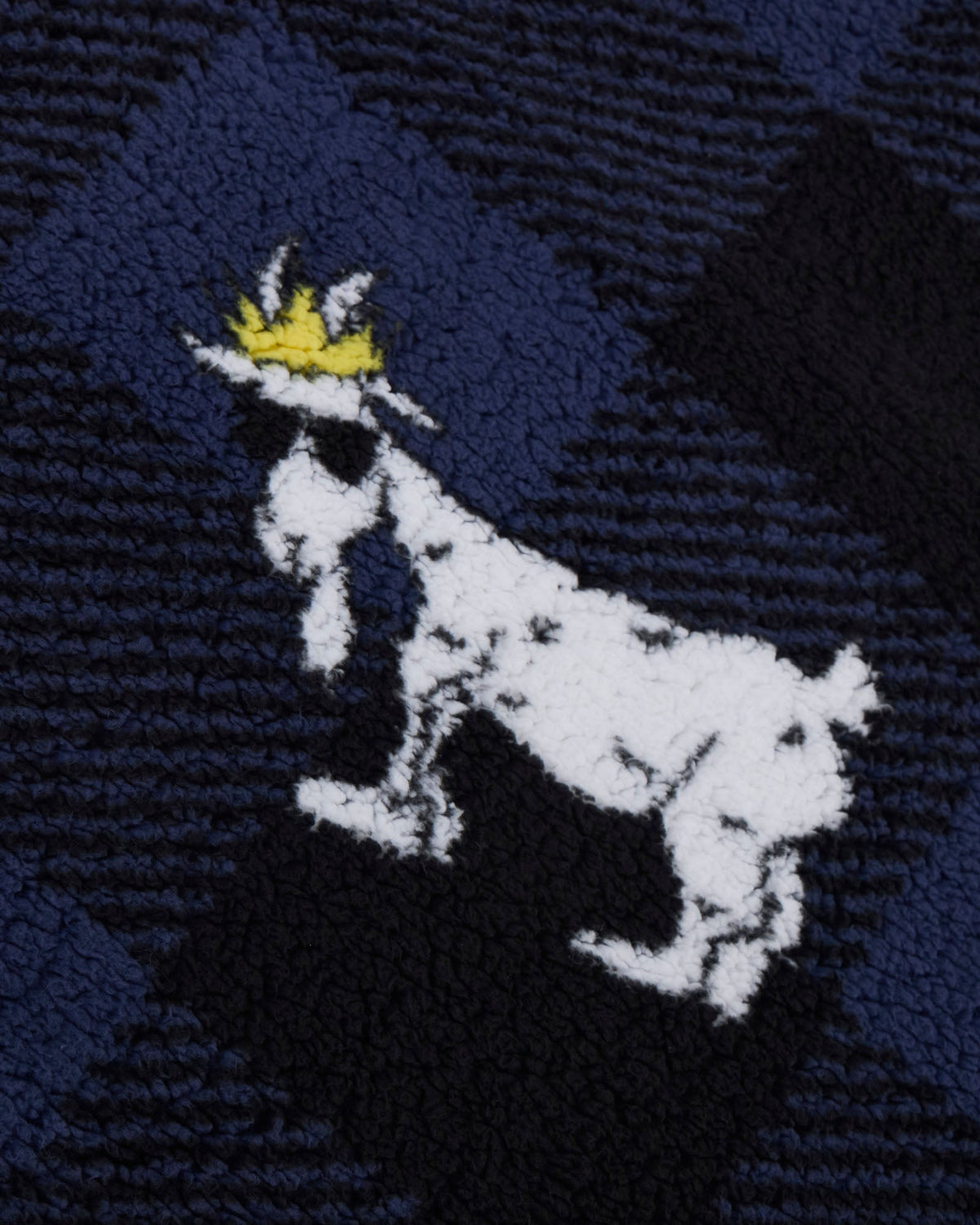 A close-up of a textured knit or fleece fabric featuring a white goat silhouette wearing a small yellow crown, set against a dark navy and black plaid background.
