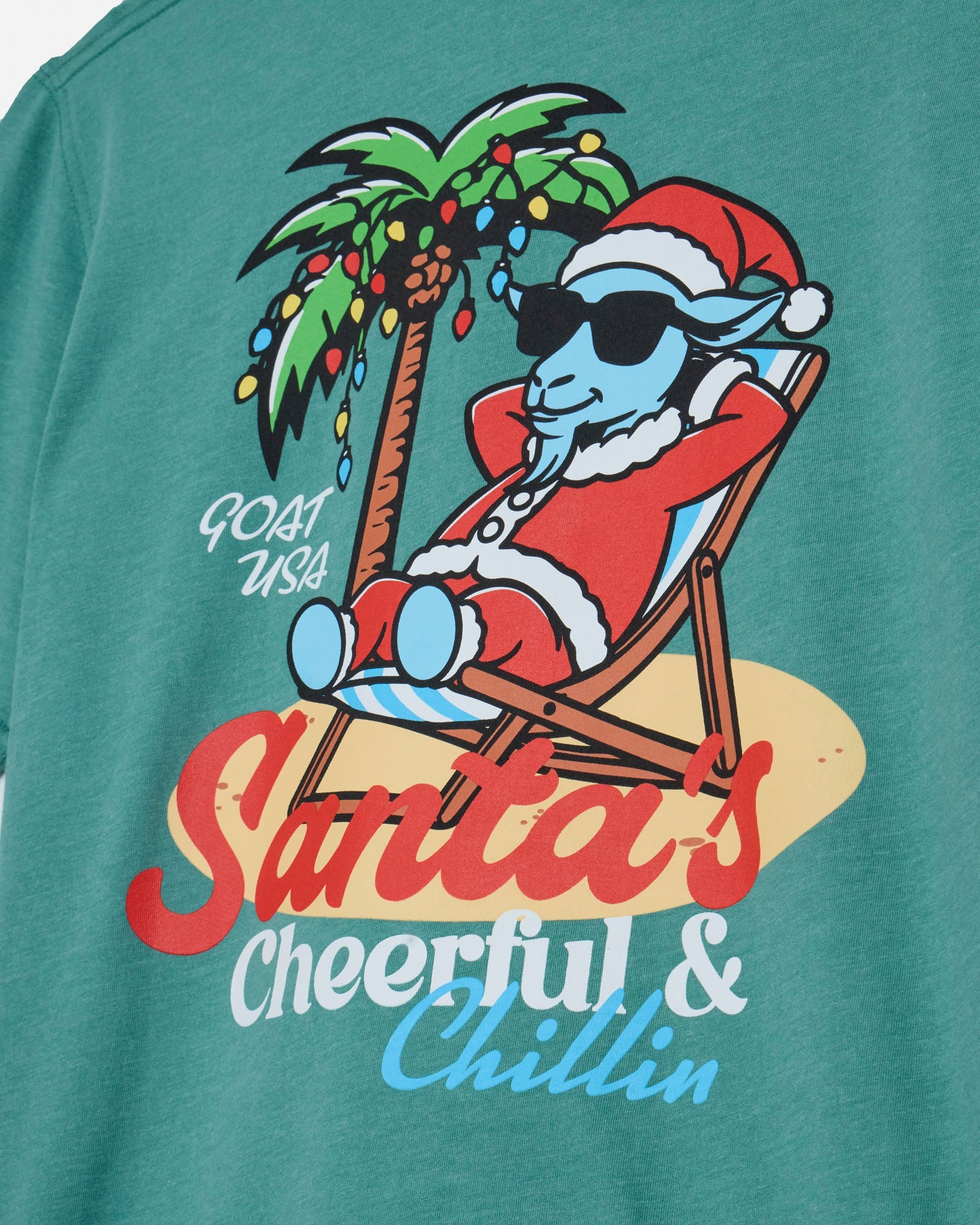 T-shirt with a graphic of a goat in Santa Claus attire relaxing under a palm tree with Christmas lights.