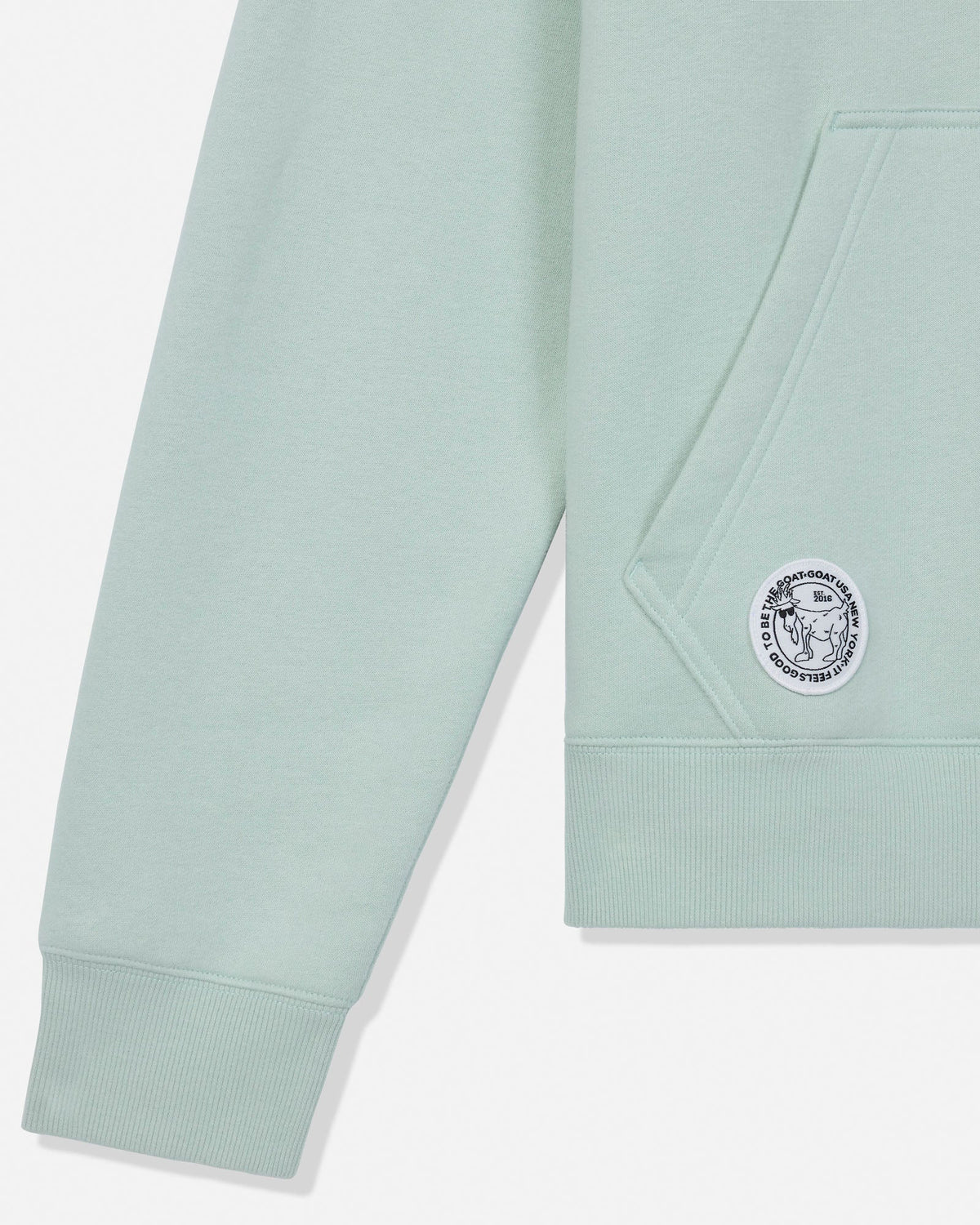 Close-up of a pale mint green sweatshirt showing the sleeve, ribbed cuff, front pocket, and a circular embroidered logo patch on the pocket.