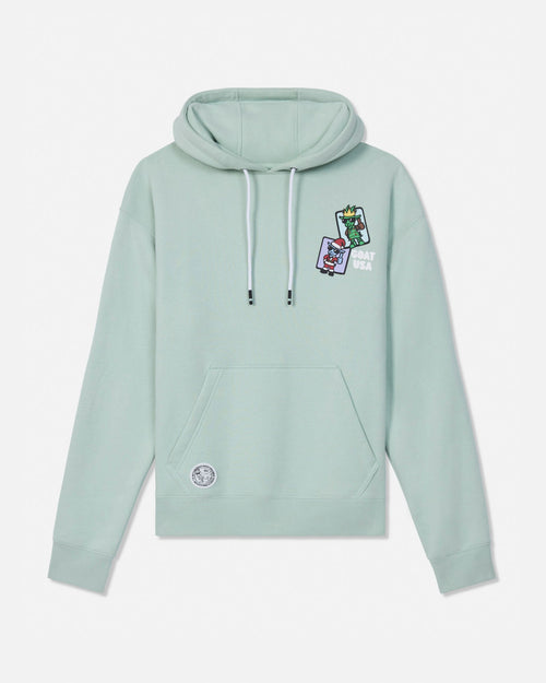 A sage green pullover hoodie with white drawstrings, featuring a small colorful graphic patch on the left chest and a circular patch on the lower left front pocket, displayed against a light gray background.