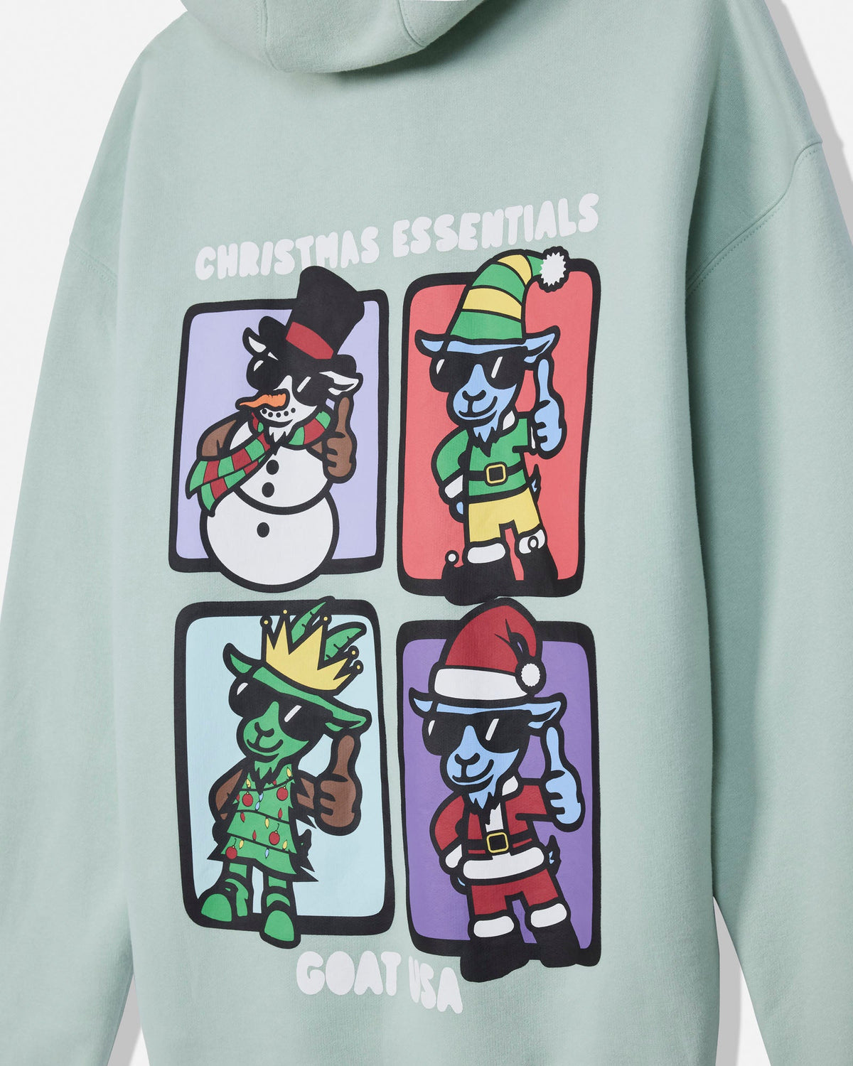 A close-up of a sage green hoodie featuring a colorful graphic print on the front. The graphic displays four cartoon Christmas characters in separate panels with the text 'CHRISTMAS ESSENTIALS' above and 'GOAT USA' below.