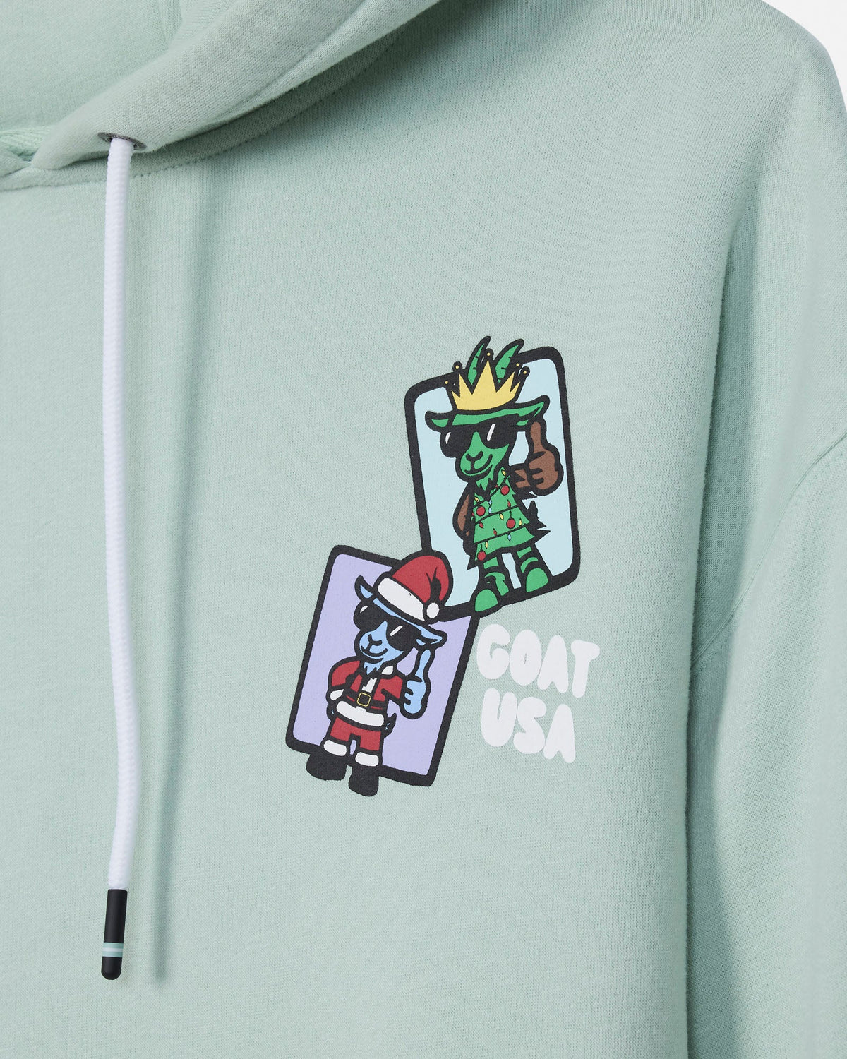 A close-up of a mint green hoodie featuring a graphic print of two cartoon turtles wearing crowns and sunglasses, one in a Santa hat, with the text 'GOAT USA' printed next to them.
