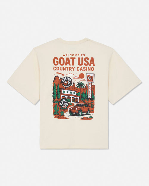 An off-white short-sleeve t-shirt with a large graphic print on the back featuring the text 'WELCOME TO GOAT USA COUNTRY CASINO' and an illustration of a red car, casino building, and desert scenery.