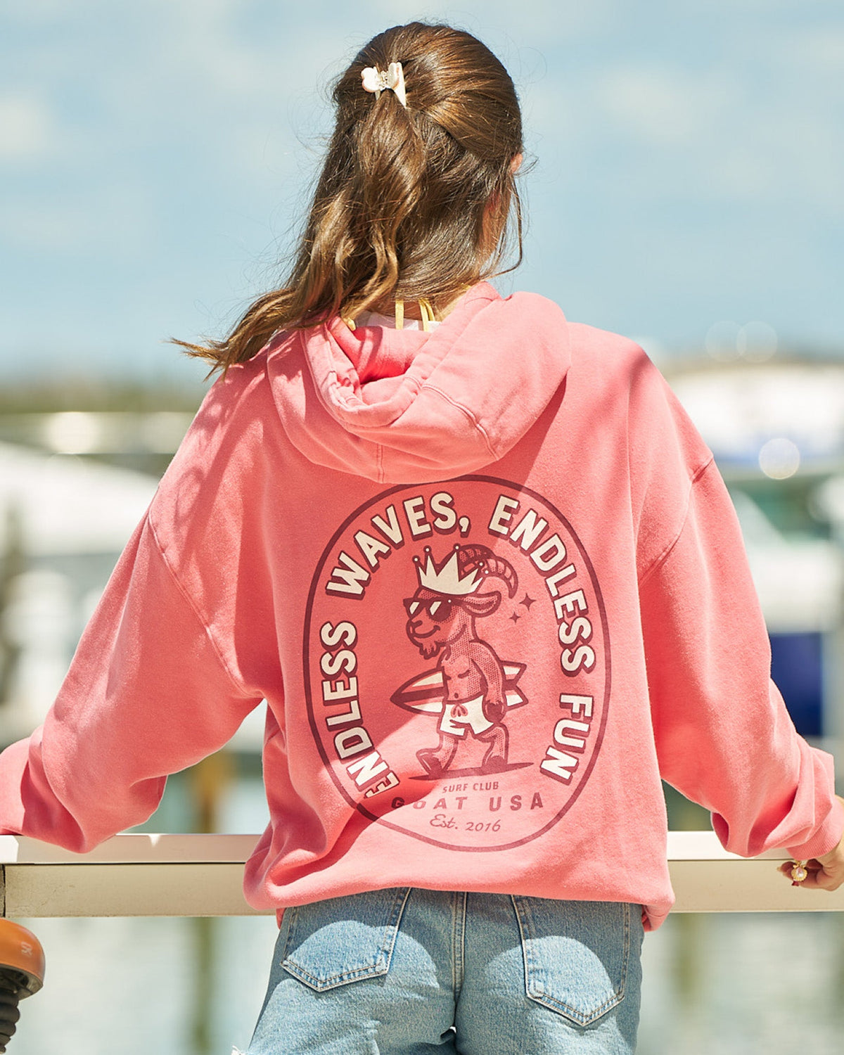 Girl in salmon-colored hoodie facing away from the camera overlooking a body of water and boats