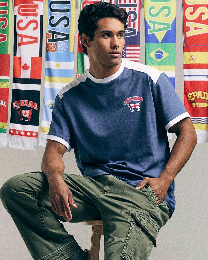 Man sitting on a stool wearing a blue t-shirt with a logo and green pants, with colorful flags in the background.