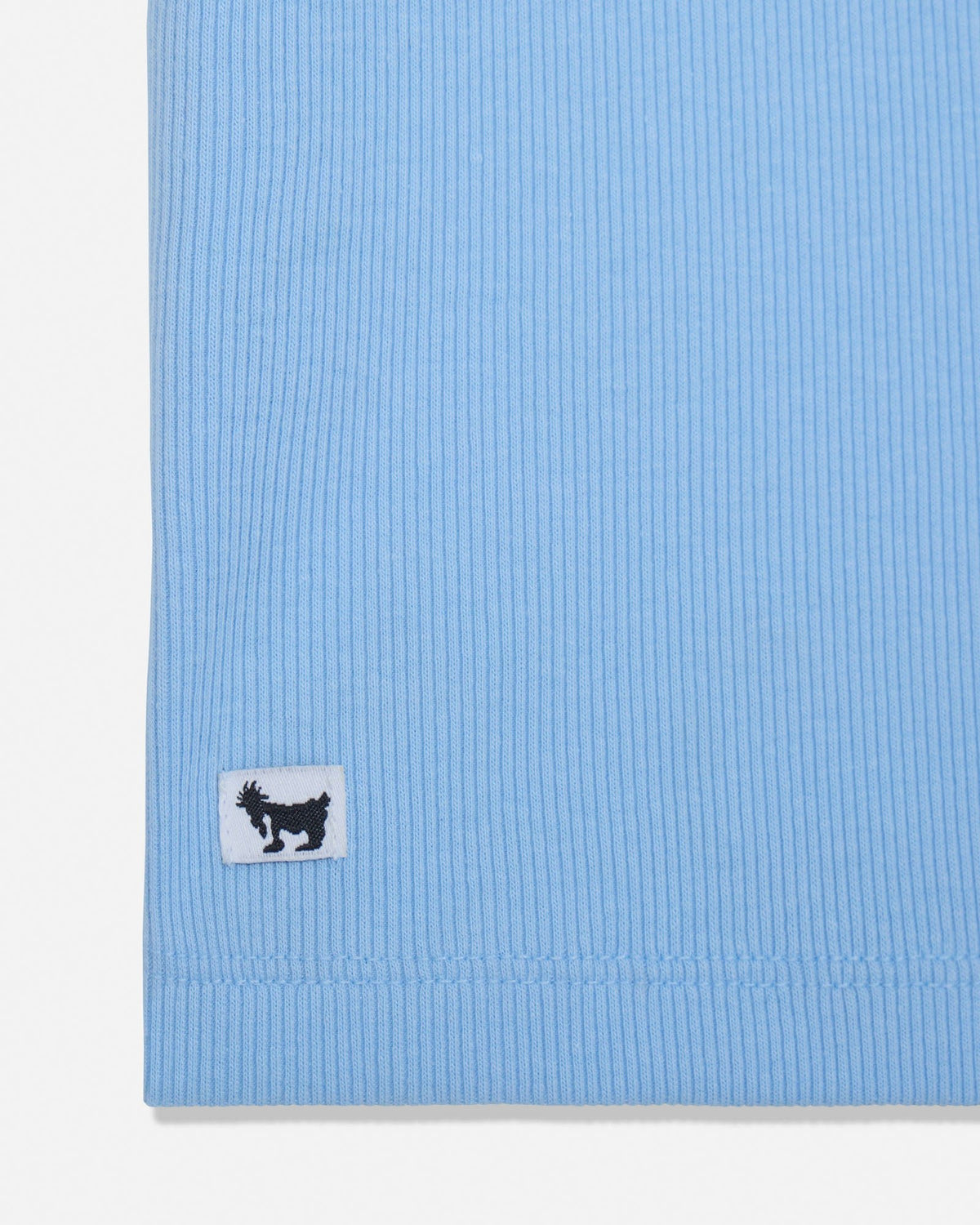 Close-up of a light blue ribbed knit garment corner with a small white rectangular patch featuring a black goat logo.