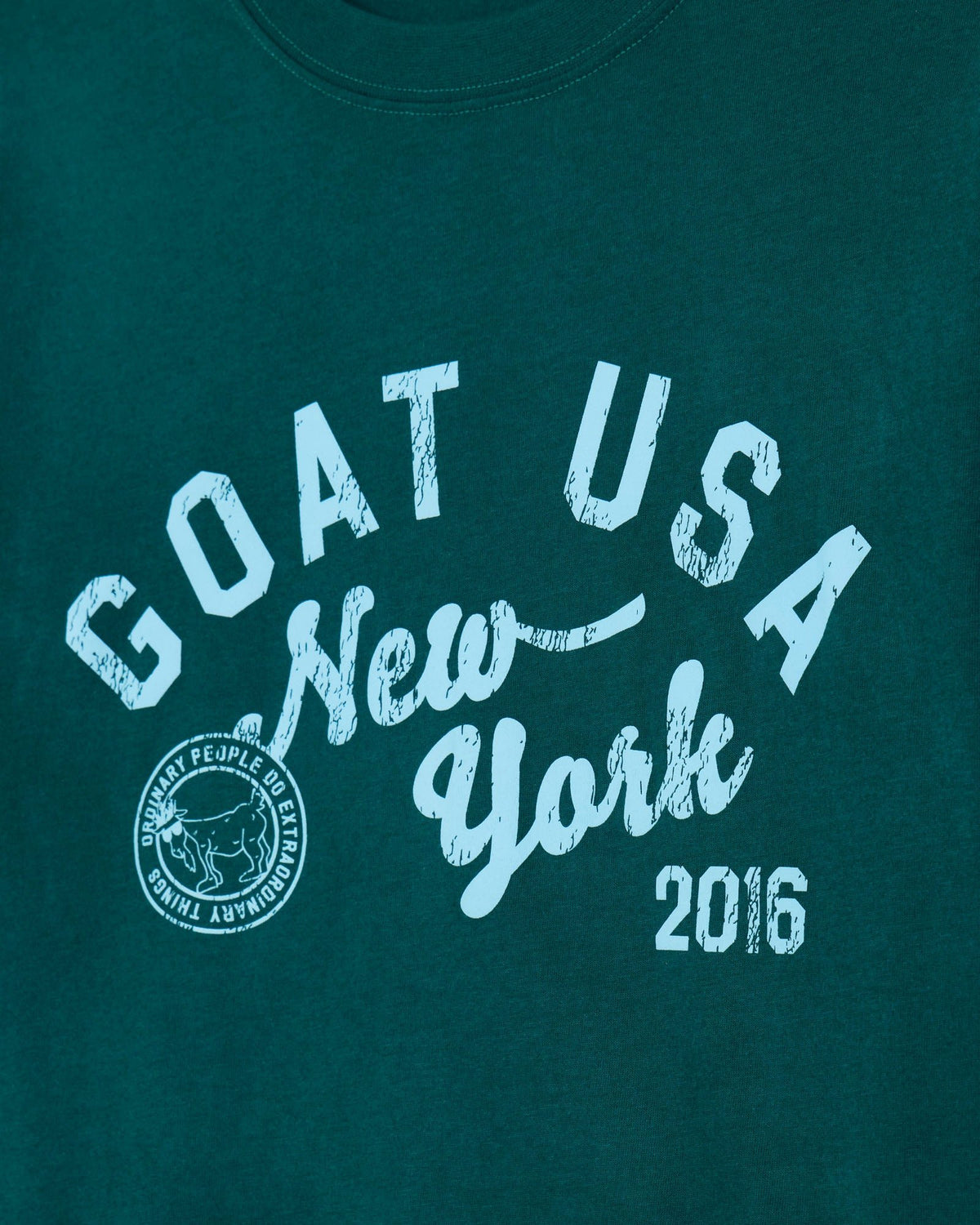 A close-up of a dark teal t-shirt featuring a distressed light blue graphic print that reads 'GOAT USA New York 2016' with a circular logo on the left side.