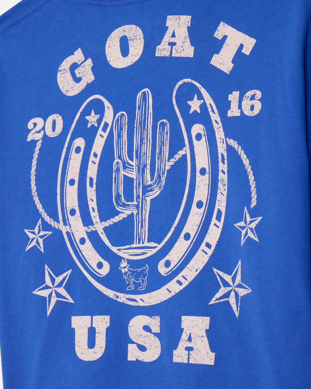 A close-up of a royal blue t-shirt featuring a distressed white graphic print with the words 'GOAT USA', stars, ropes, and a central design of a cactus growing out of a horseshoe.