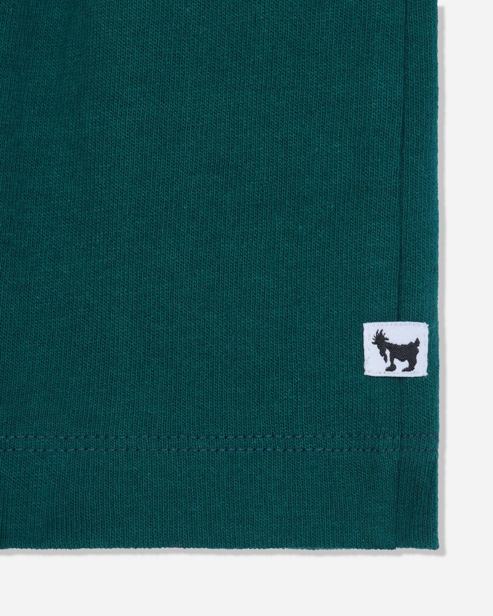 Close-up of a dark teal green knit fabric corner with a small white square patch featuring a black goat logo.