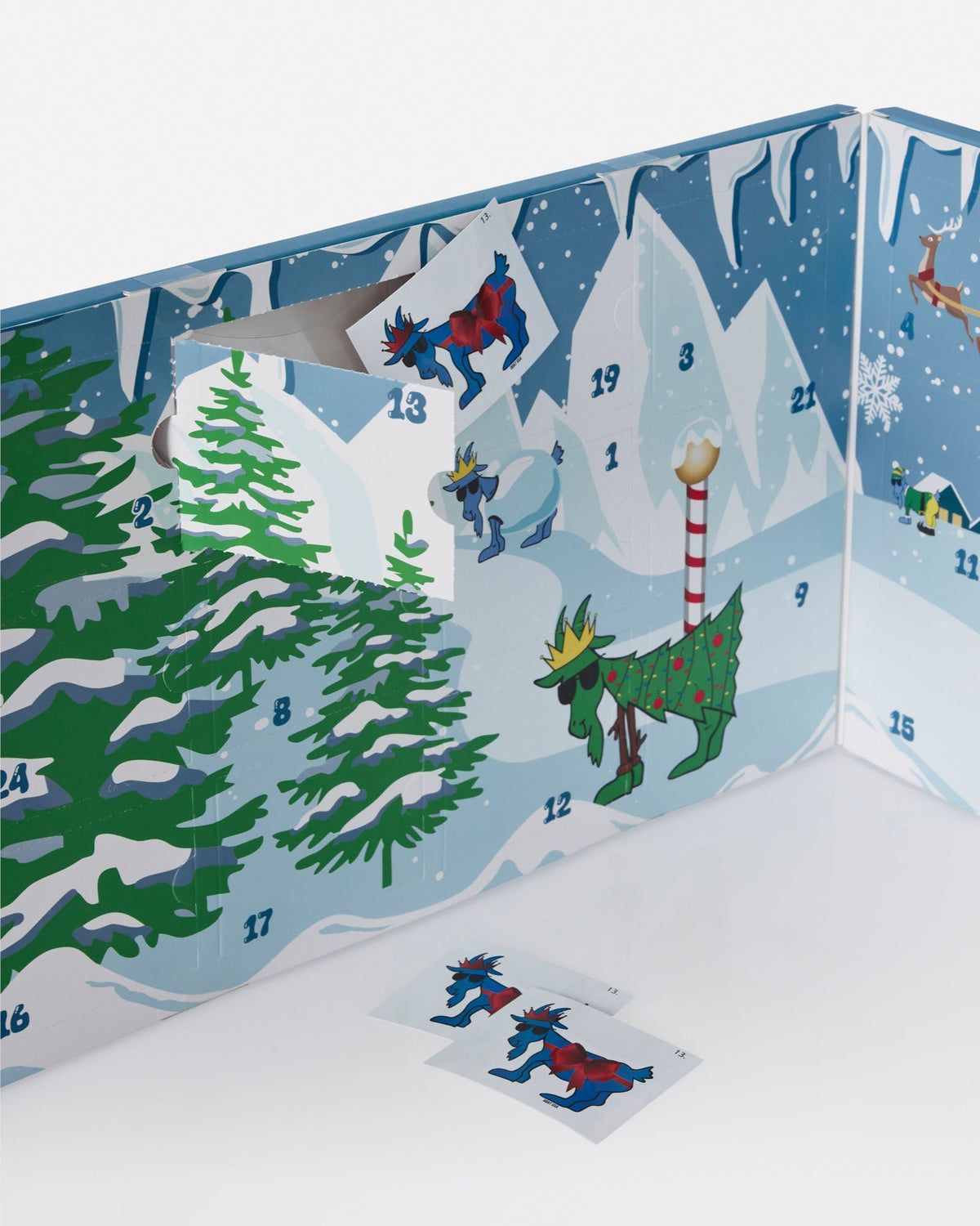 A colorful Advent calendar box with illustrated winter scenes, including snowy mountains, evergreen trees, reindeer, and numbered door openings. Two small cards with reindeer illustrations are placed in front on a white surface.