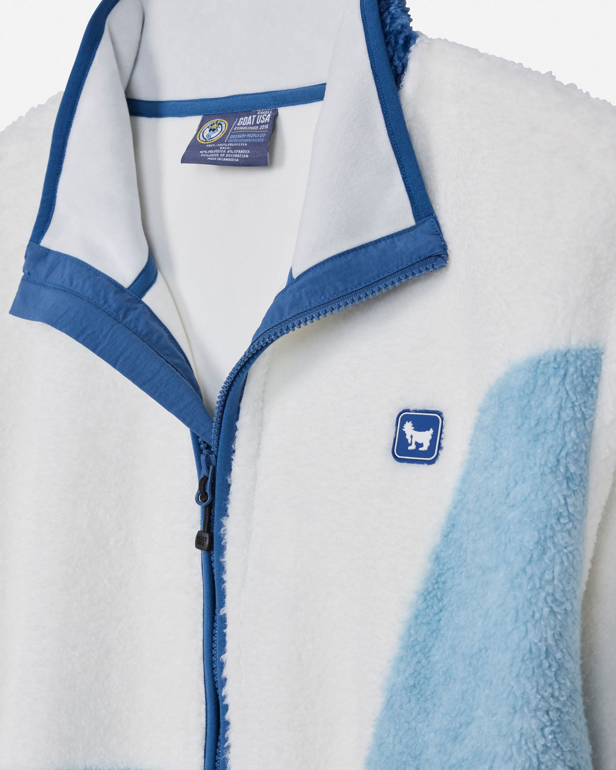 Close-up of a fleece zip-up jacket with a white sherpa-like body, blue trim, a blue zipper, and a small blue chest patch with a white goat silhouette. A fabric label is visible inside the collar.