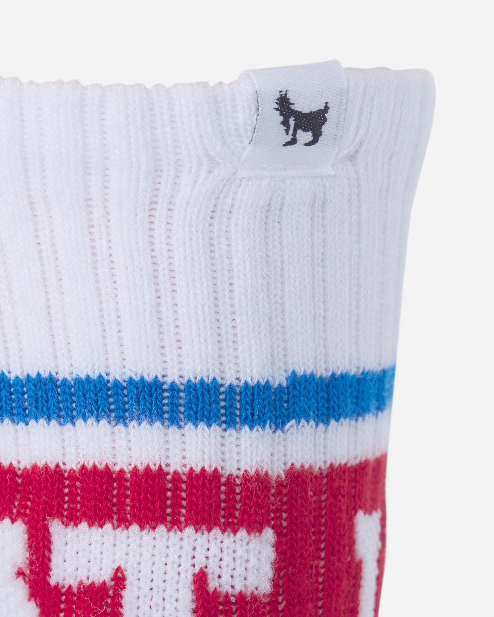 Close-up of a white ribbed knit sock with horizontal blue and red stripes and a small black goat silhouette logo on the cuff.#color_true-red