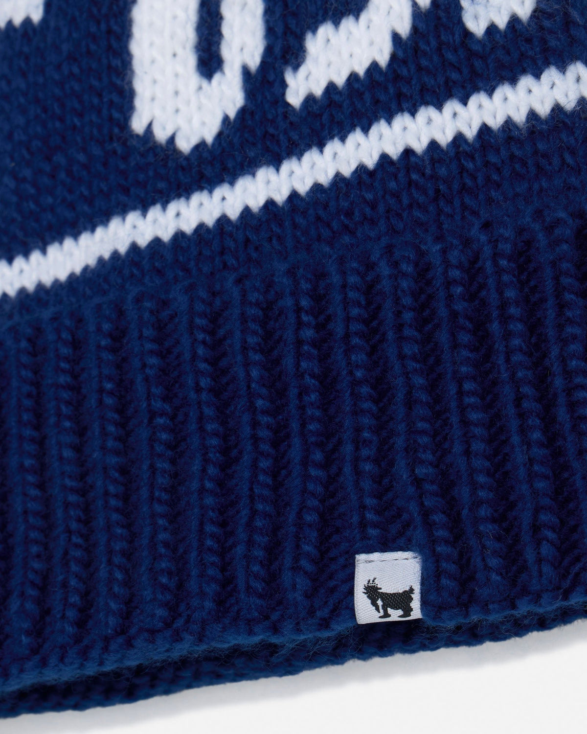 Close-up of a navy blue knit beanie with white knit lettering and a ribbed cuff, featuring a small white square tag with a black goat logo near the bottom.