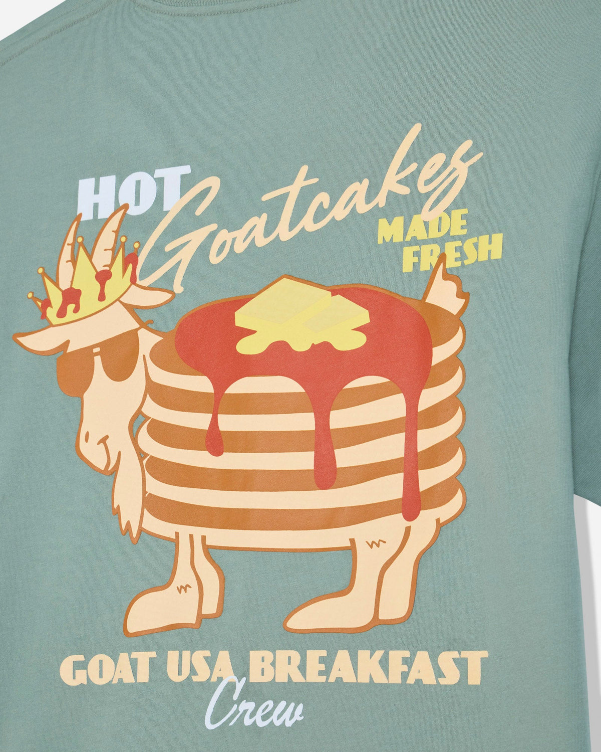 A close-up of a sage green t-shirt featuring a graphic of a goat wearing sunglasses and a crown, with a stack of pancakes topped with syrup and butter. The text reads 'HOT Goatcakes MADE FRESH' and 'GOAT USA BREAKFAST Crew'.