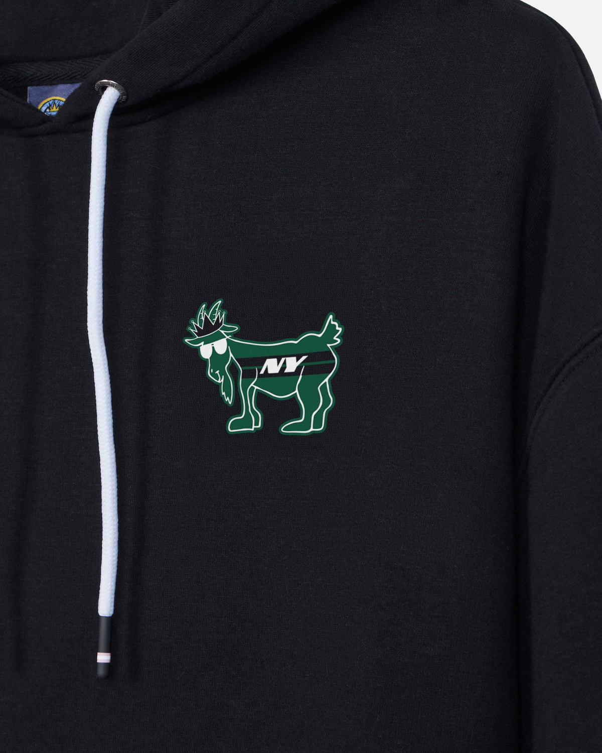 Close-up of a black pullover hoodie featuring a green embroidered goat graphic with sunglasses and the letters 'NY' on the left chest, with a white drawstring.