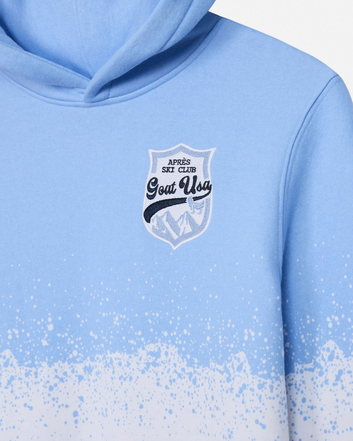 A close-up of a light blue hoodie with a white and navy graphic shield on the chest reading 'Après Ski Club Goat Usa' and a paint-splatter gradient at the bottom hem.